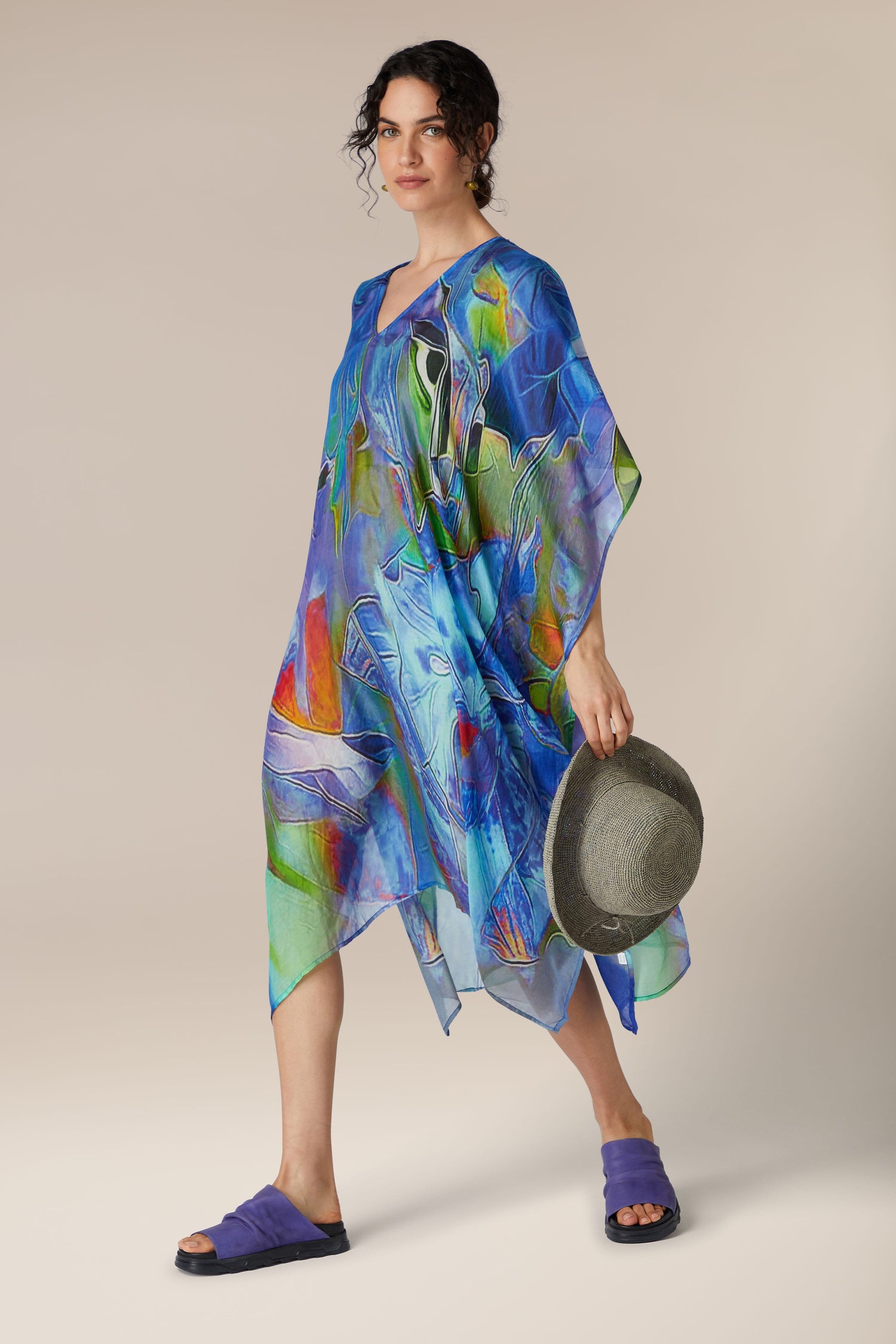 A woman in a flowing, colorful Tiffany Lamp Silk V Neck Kaftan holding a gray hat, posing against a neutral background.