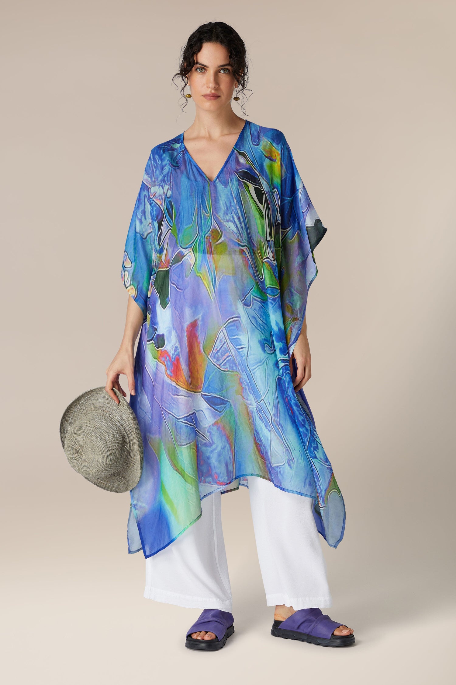 A woman holding a hat stands wearing a colorful blue-toned Tiffany Lamp Silk V Neck Kaftan over white trousers, paired with navy sandals.