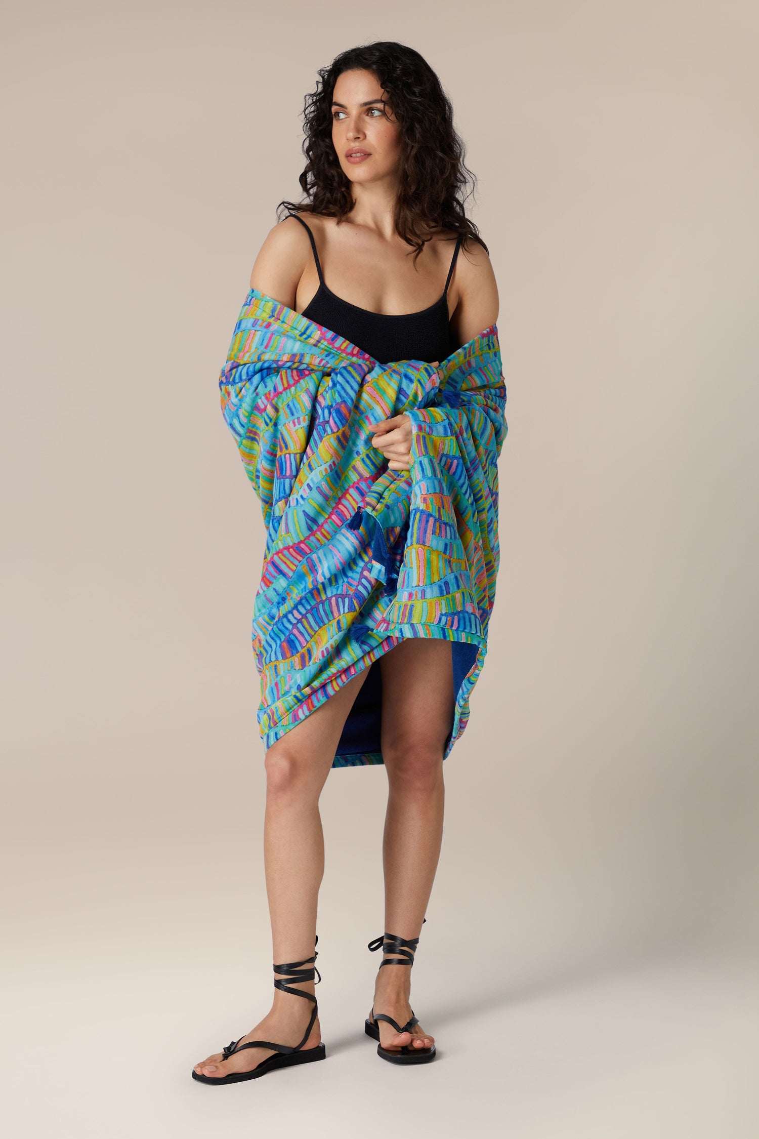 A woman standing while holding a colorful patterned Rainbow Wave Beach Towel, looking to the side.