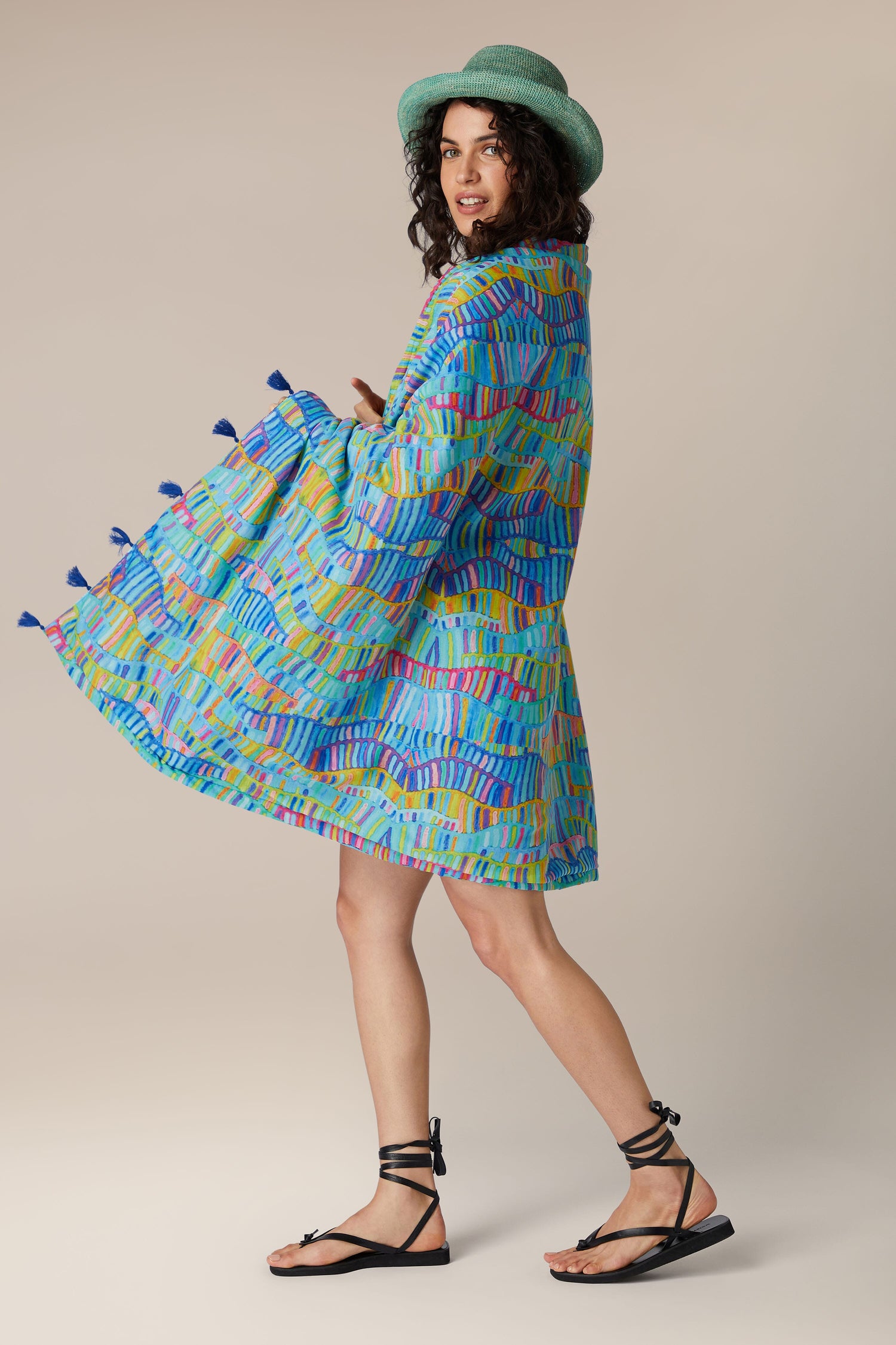 A woman in a vibrant blue patterned coat and holding a Rainbow Wave Beach Towel is walking and looking over her shoulder.