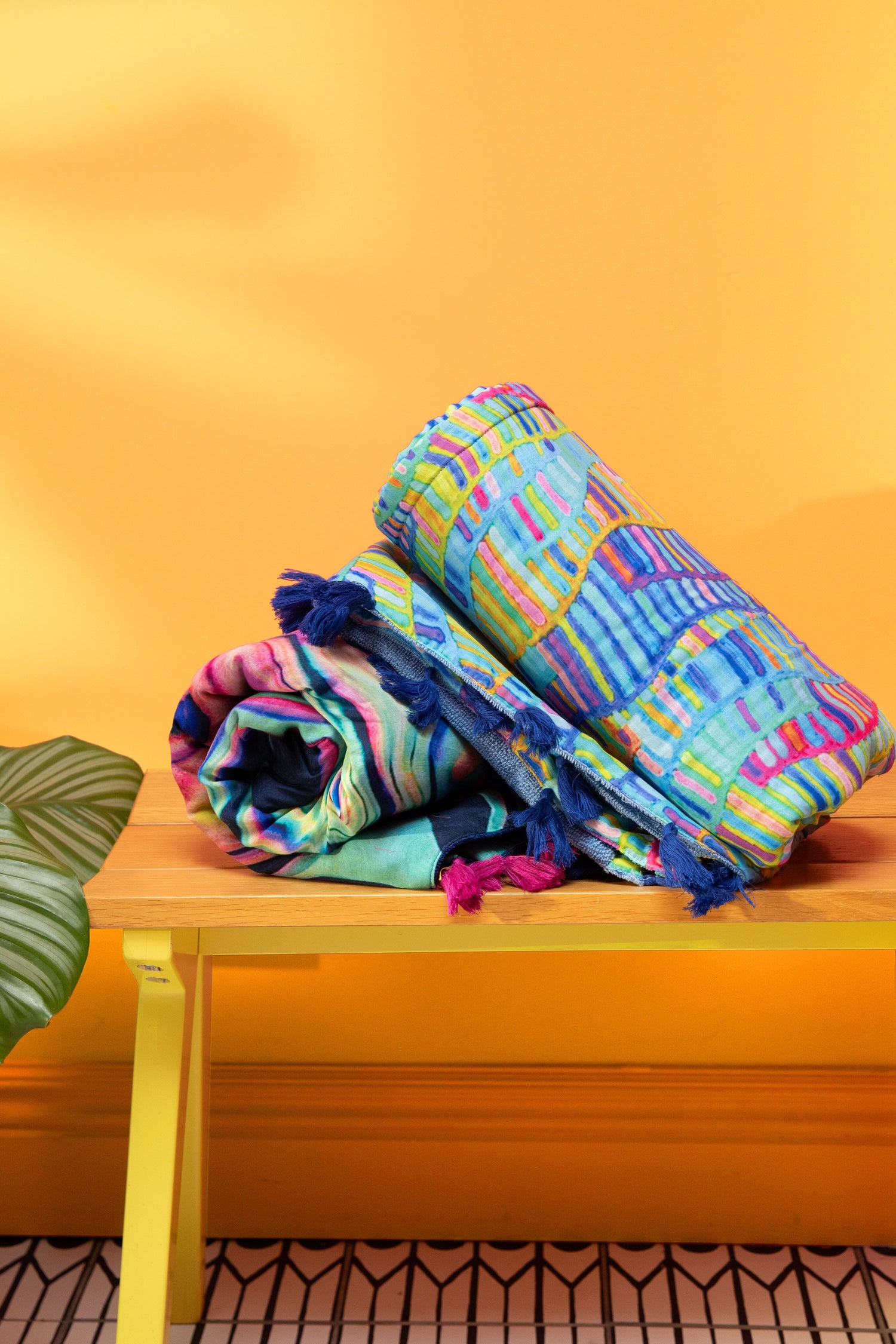 Two colorful, patterned blankets, including the vibrant Rainbow Wave Beach Towel, are neatly rolled and stacked on a wooden bench against a bright orange wall. Perfect as summer beach essentials, these pure cotton accents bring both comfort and style. A green leaf is partially visible on the left side of the image.