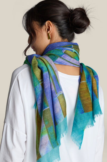 A person with dark hair in a bun is wearing a white top and a vibrant Fragment Wool Scarf featuring blue, green, and yellow patterns. Made in India from premium wool, the scarf adds an eye-catching touch as the person is shown from the back.