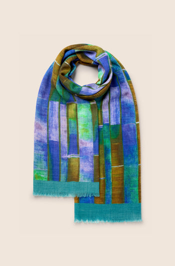 The Fragment Wool Scarf is a rectangular, multicolored piece made in India, showcasing abstract patterns in green, blue, purple, and brown with frayed edges on a light background. This premium wool scarf seamlessly combines style and comfort.