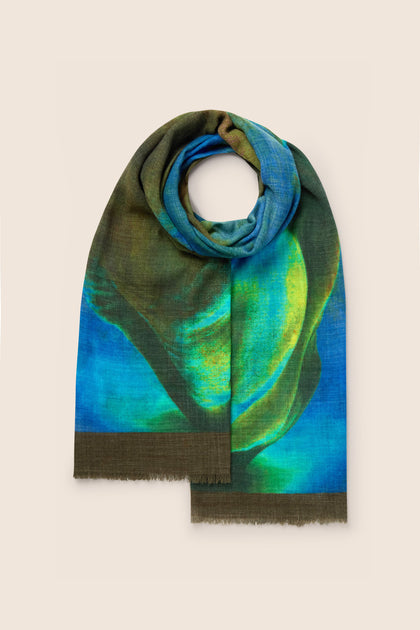 A folded Ocean Deep Wool Scarf, displaying a vibrant and multicolored abstract pattern in shades of blue, green, and brown against a plain, light-colored background. This premium wool accessory boasts exquisite sea-tone colors that elevate any ensemble.