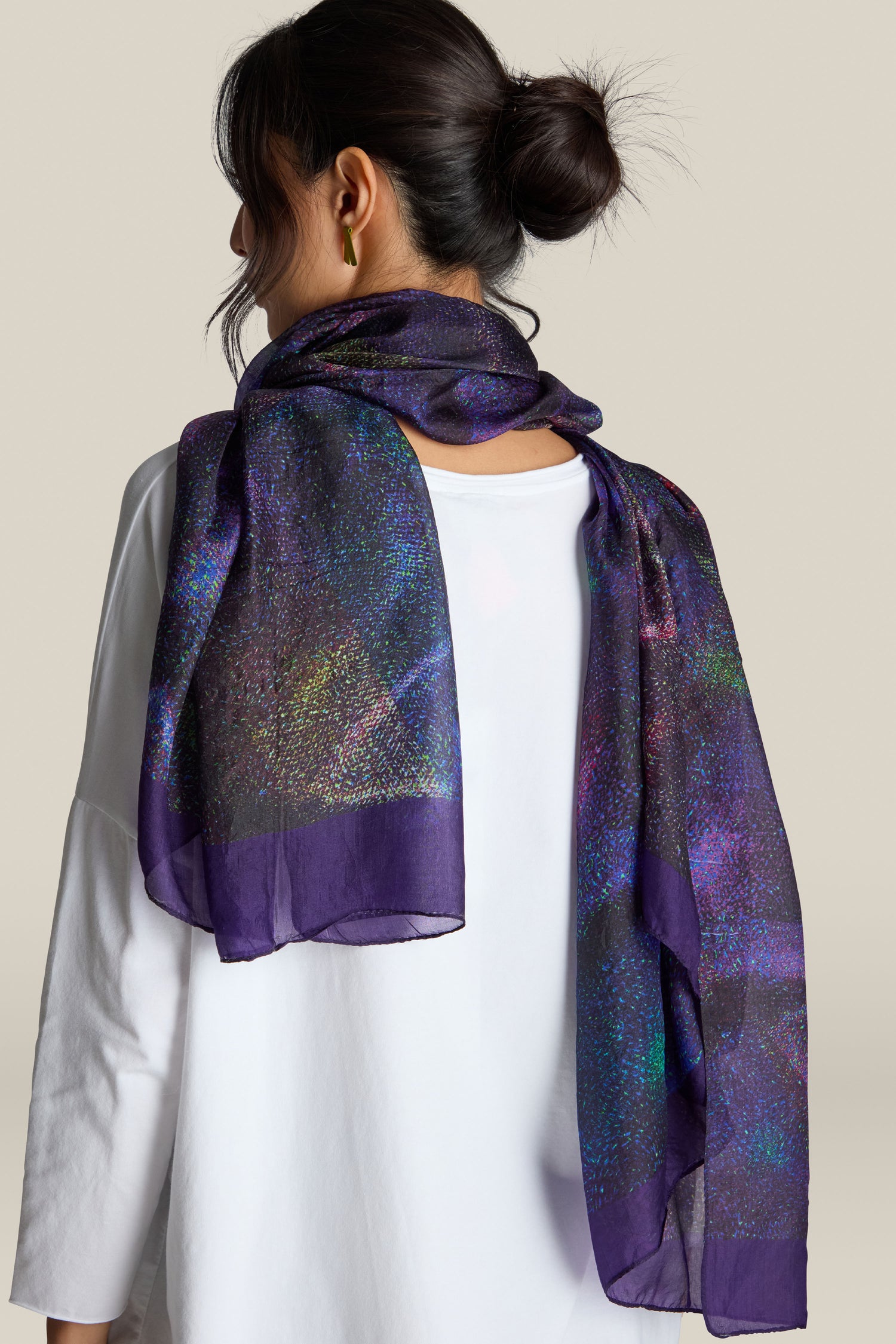 Woman with dark hair in a bun, wearing a white top and draped over her shoulders is the luxurious Cosmos Silk Scarf, its vibrant and glittery purple fabric catching the light as she is viewed from the back.