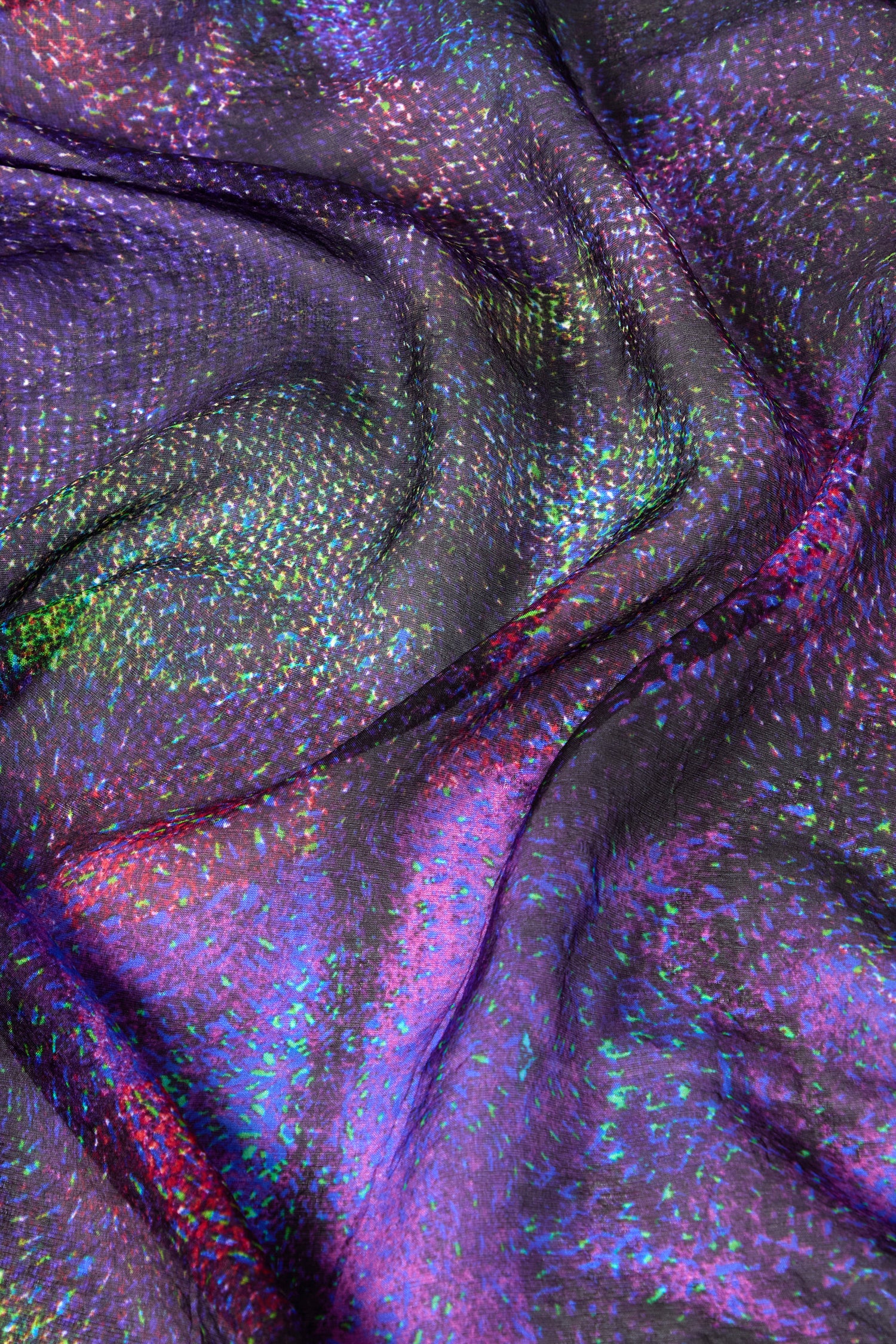 Close-up of iridescent fabric with a shimmering, multicolored pattern. The luxurious Cosmos Silk Scarf has a glossy surface and exhibits hues of purple, green, blue, and pink.