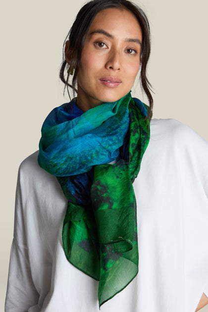 A person with long hair wears a white top and a Galaxy Silk Scarf, featuring shades of green and blue, standing against a plain background.