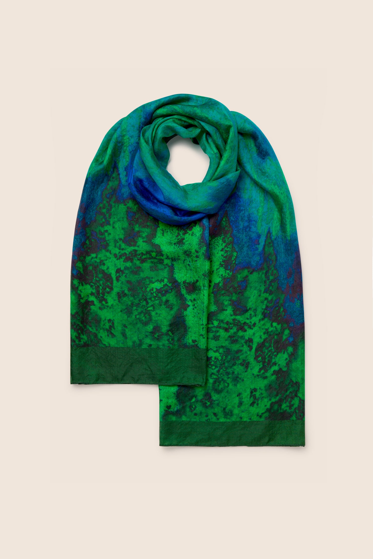 A Galaxy Silk Scarf, featuring a captivating blue and dark green abstract forest pattern, is laid flat on a plain background. This celestial accessory in soothing blue and green tones adds an elegant touch to any outfit.