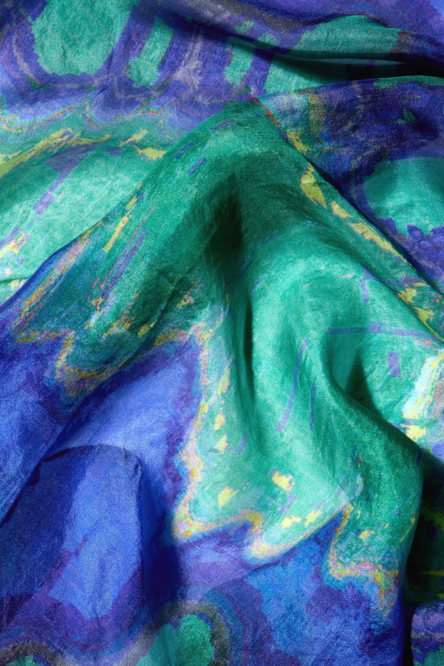 A close-up of the Mineral Silk Scarf reveals crumpled fabric in shades of blue and green, adorned with a wavy, abstract pattern. The texture and vibrant colors create a dynamic visual effect, showcasing its refined luxury.