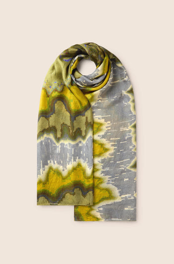 The Mineral Silk Scarf showcases expert craftsmanship, presenting a folded shawl adorned with an abstract pattern in hues of green, yellow, and gray, set against a plain beige backdrop.