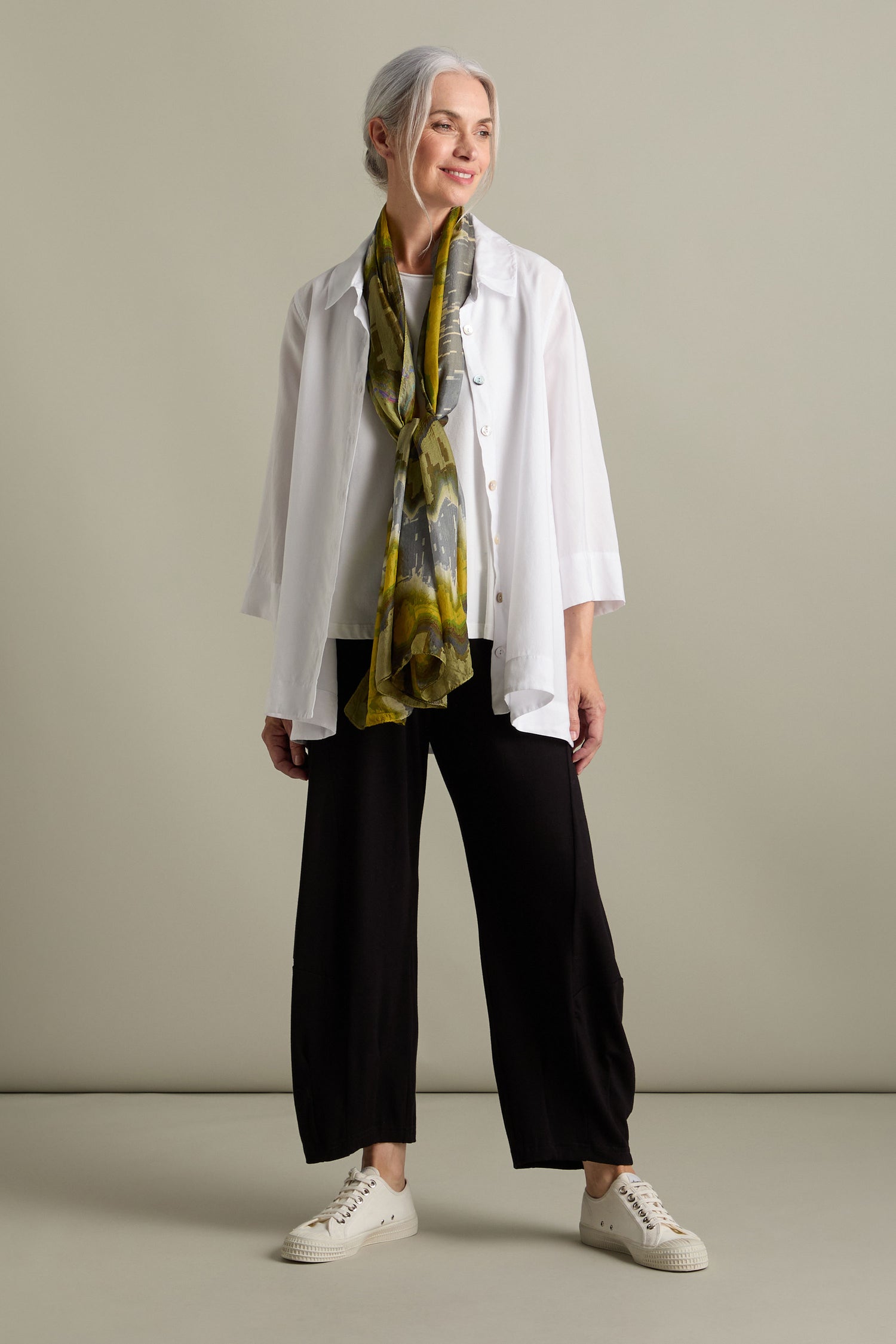 An older woman with gray hair, dressed in a white shirt, black pants, and white sneakers, stands against a plain background. She wears the Mineral Silk Scarf, exuding refined luxury as she looks to the side and smiles.