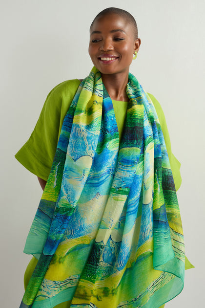 Wearing a green outfit and the vibrant Painted Ocean Silk Scarf, a person stands against a light backdrop, smiling to the right.