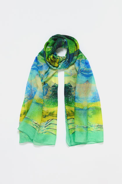 The Painted Ocean Silk Scarf, inspired by the ocean, features a wavy landscape pattern in vibrant shades of green, blue, and yellow draped elegantly against a white background.