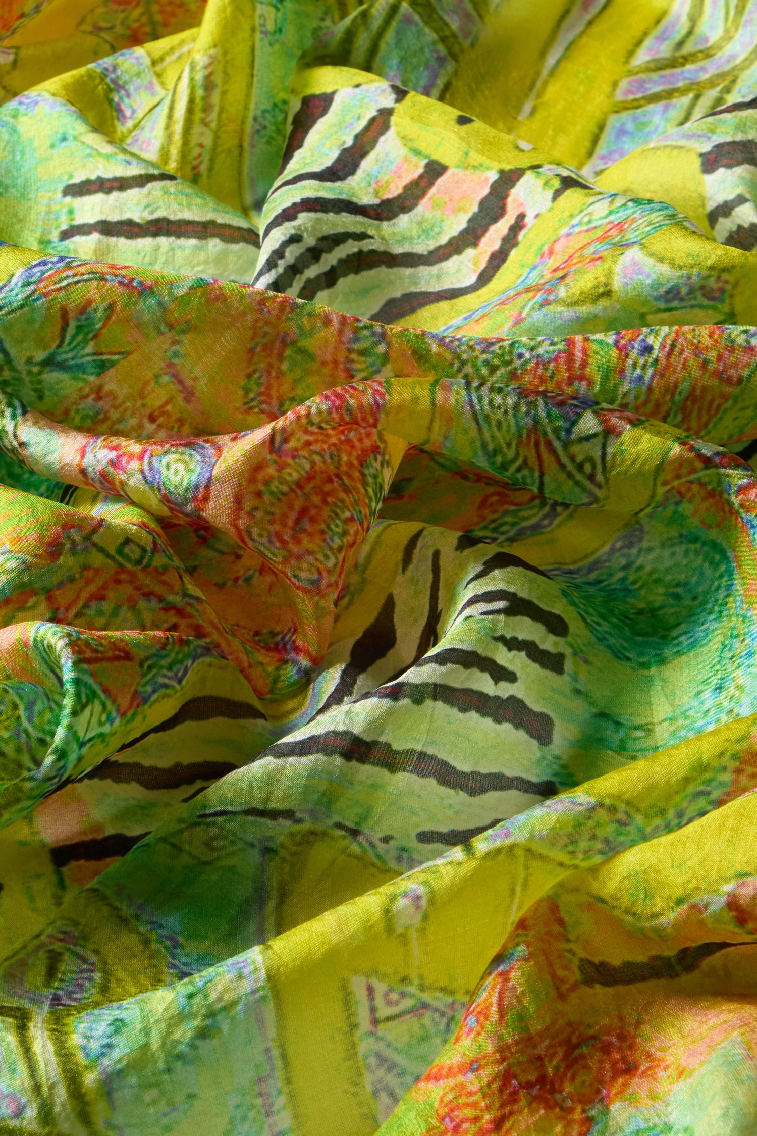 A close-up of the Citrine Silk Scarf reveals its vibrant abstract patterns in green, orange, and black. The scarf's soft, wavy texture highlights a blend of stripes and exotic circular motifs.