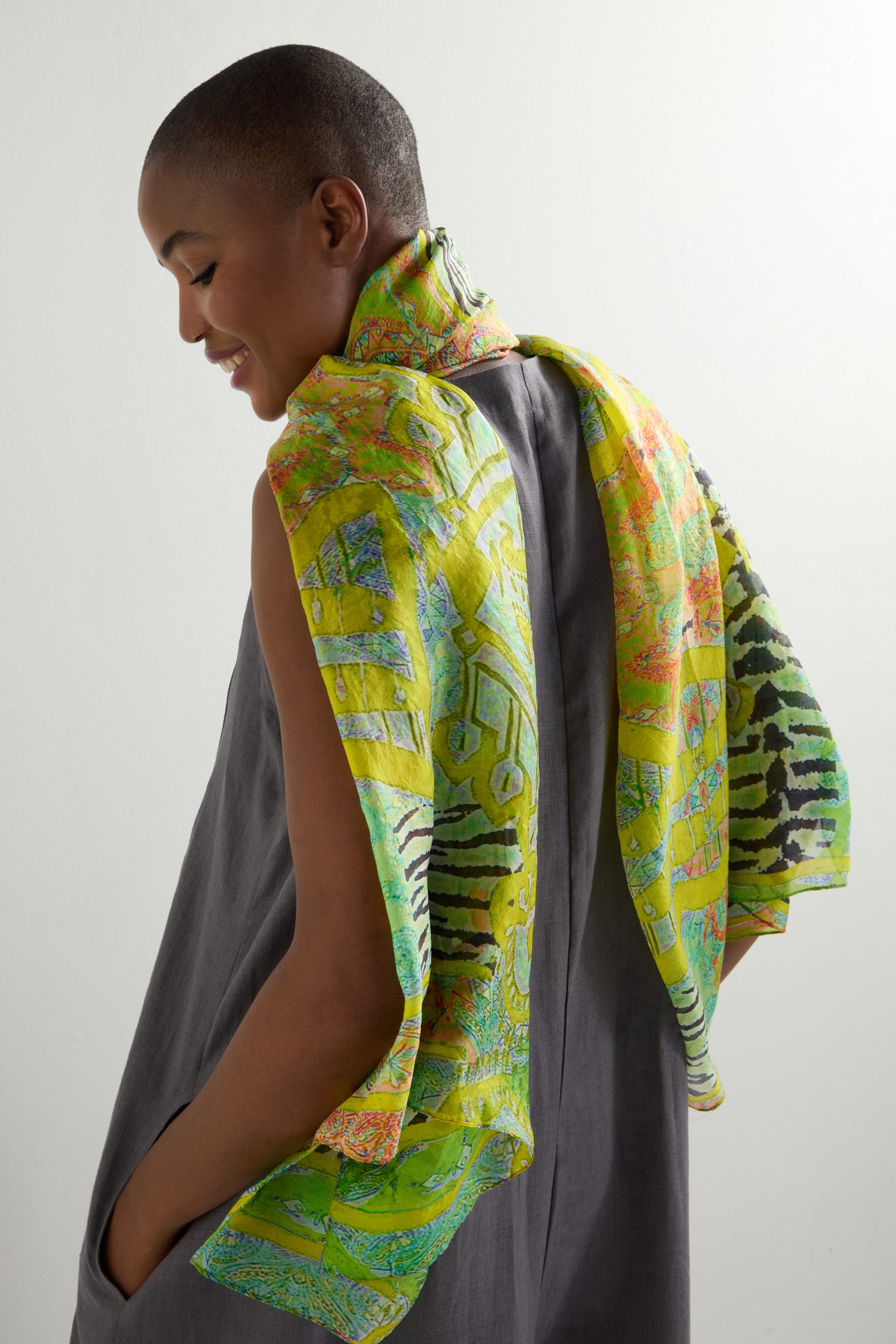 Wearing a gray sleeveless dress, a person has a Citrine Silk Scarf with exotic motifs draped over their shoulders. They look downward against a neutral background, with the vibrant scarf adding an intriguing touch.