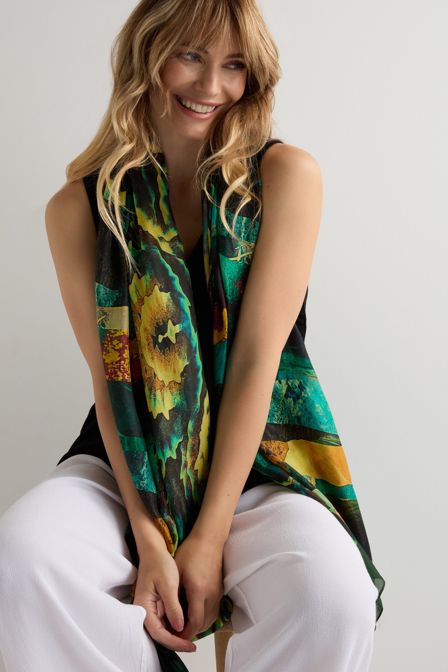 A smiling woman with long blonde hair sits on a stool, wearing a black sleeveless top, white pants, and the Northern Lights Silk Scarf, which features a luxurious green and yellow abstract design.