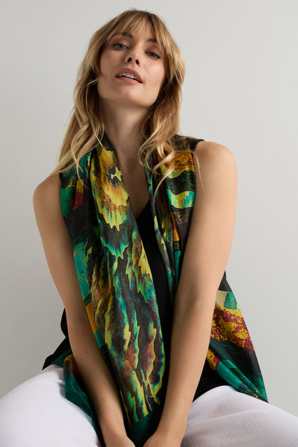 A woman with blonde hair wears the Northern Lights Silk Scarf, featuring green and yellow abstract designs, over a black top and white pants against a plain light background.