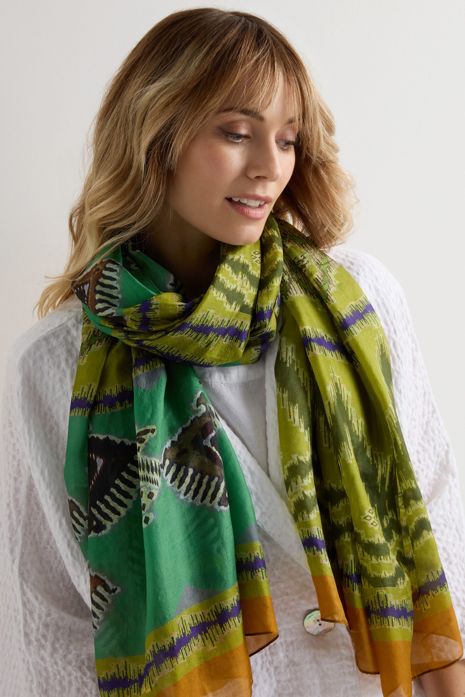 A woman in a white textured top smiles slightly downward, wearing the Aztec Silk Scarf featuring green patterns, orange edges, and zebra designs.