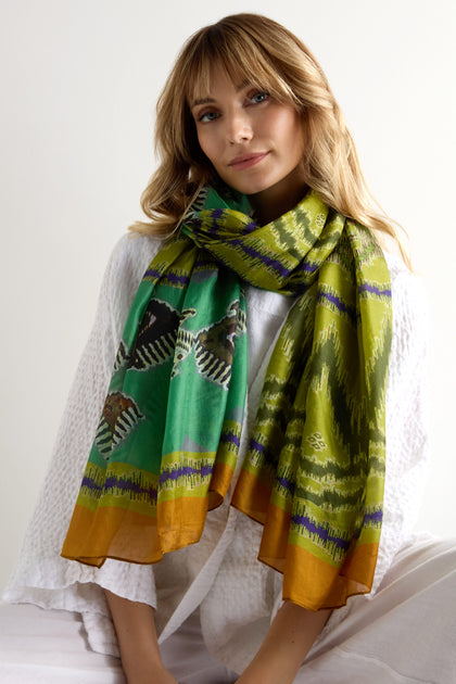Woman wearing a white textured top and the Aztec Silk Scarf—green silk with yellow, black, purple, and orange tribal-inspired patterns—sits against a plain light background.