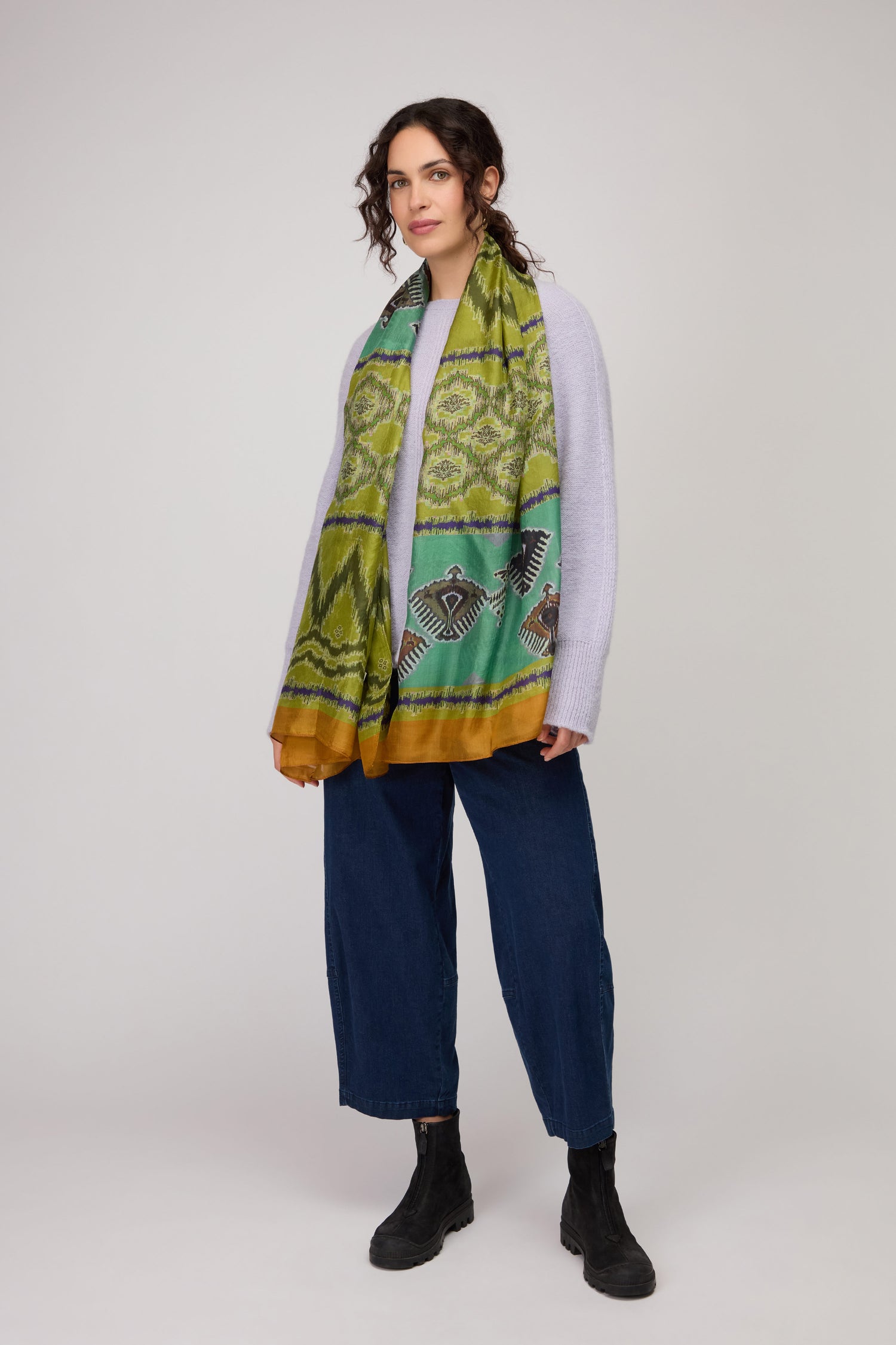 A woman wears a light gray sweater, blue wide-leg pants, black boots, and the Aztec Silk Scarf—a green patterned scarf with bird designs—against a plain light background. The Aztec Silk Scarf adds a unique touch to her stylish look.