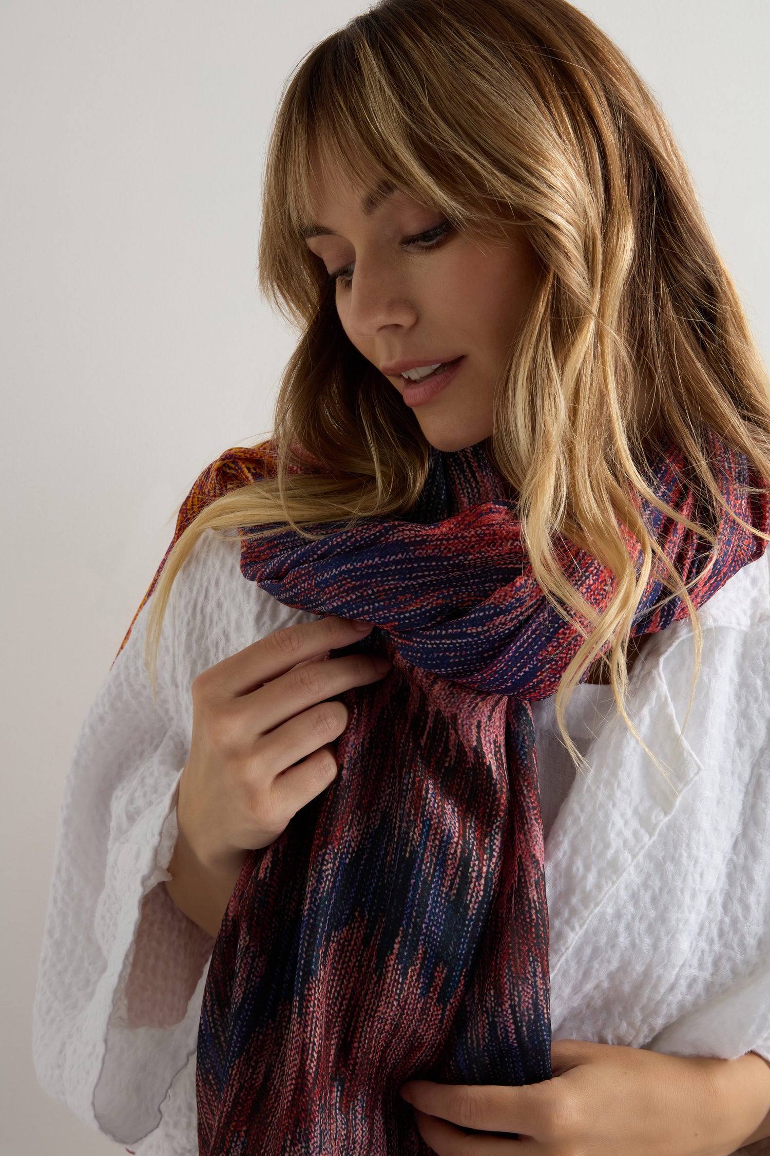 A woman in a white textured robe looks down, touching the Shaded Sketch Silk Scarf—its multicolored lightweight fabric standing out against a plain background.