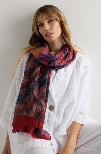 A person wears a textured white button-up top and loose white pants, styled with the Shaded Sketch Silk Scarf draped around their neck, seated against a plain light background.