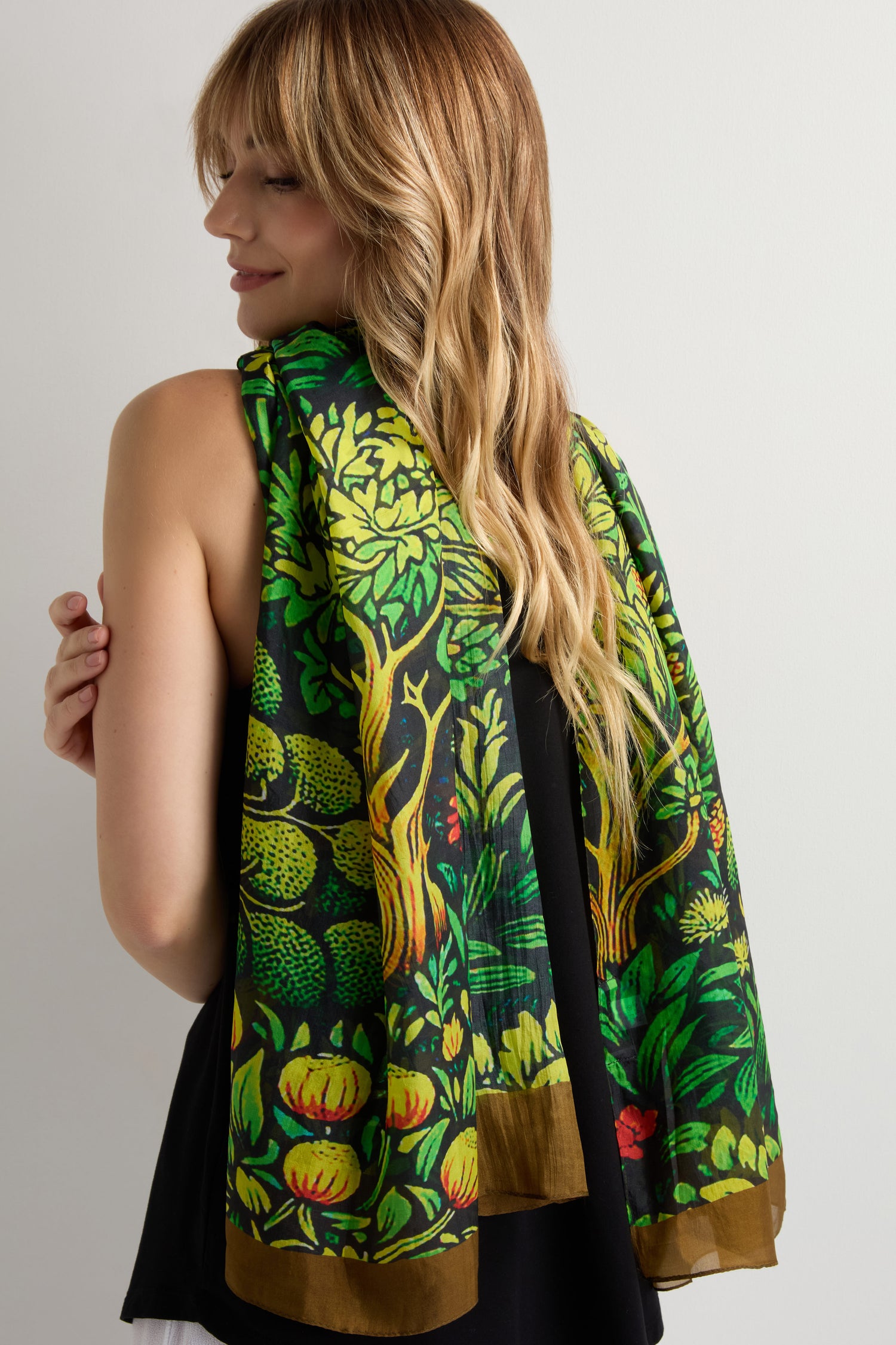A woman with long blonde hair, facing away from the camera, wears a black sleeveless top and drapes the Autumnal Garden Silk Scarf—featuring green, yellow, and brown patterns—over her shoulders.