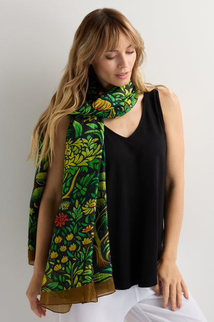 A woman stands against a light-colored wall wearing a black sleeveless top, white pants, and the Autumnal Garden Silk Scarf with its colorful floral design draped around her neck.