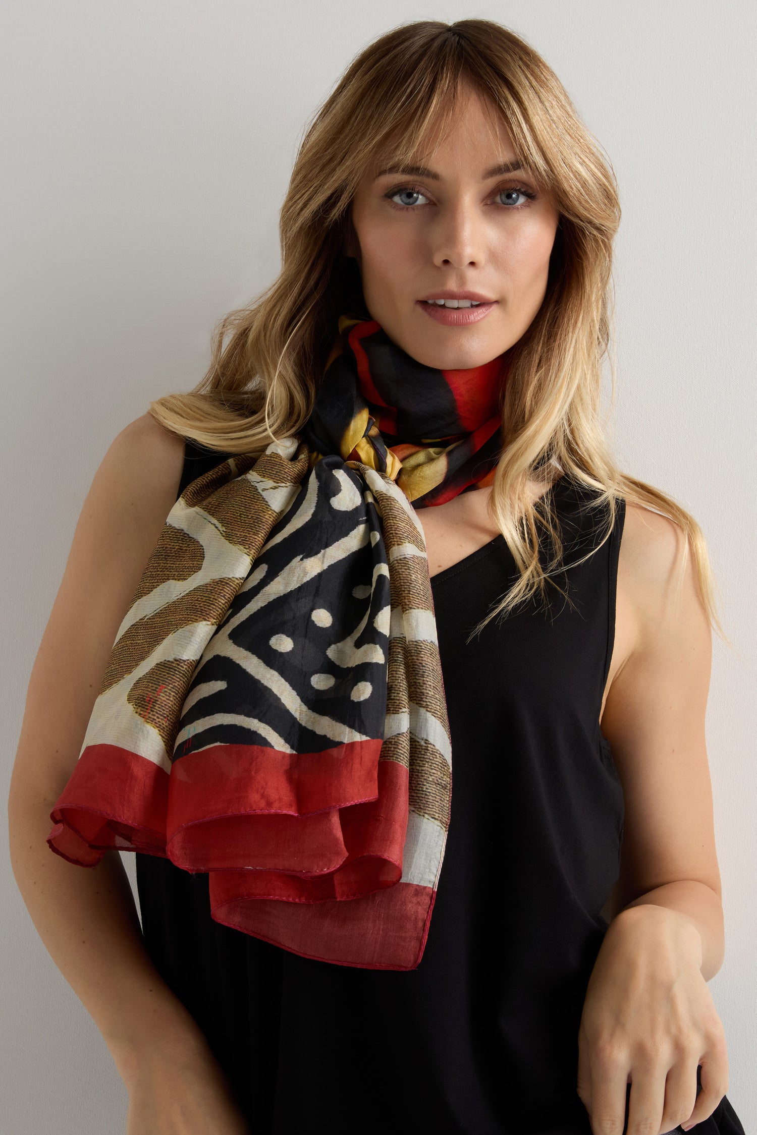 A woman with long blonde hair stands against a neutral background, wearing a black sleeveless top and the Ancient Silk Scarf—a modern statement piece with red, black, white, and gold tribal-inspired accents.