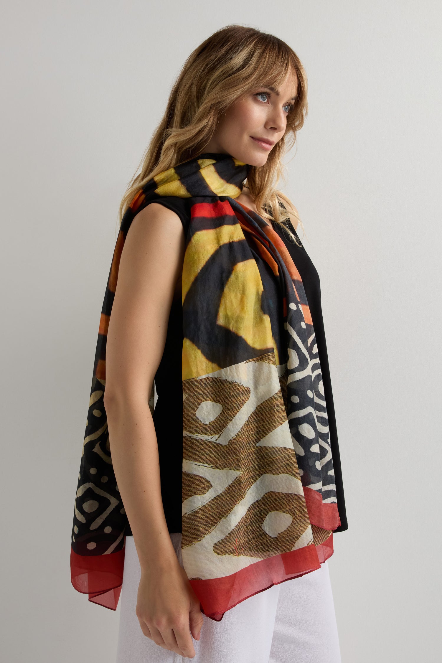A woman in a sleeveless black top and white pants stands against a plain background, wearing the Ancient Silk Scarf—a modern statement piece with a red, yellow, black, and white tribal-inspired print.