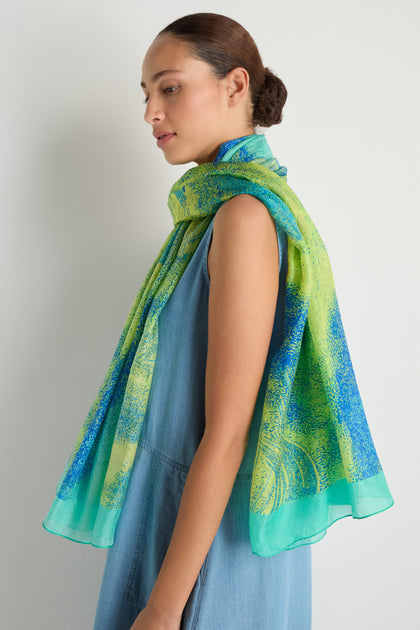 A woman stands in profile against a white background, wearing the Sky Dance Silk Scarf—a blue and green lightweight accessory with a fluid drape—over a sleeveless denim dress.
