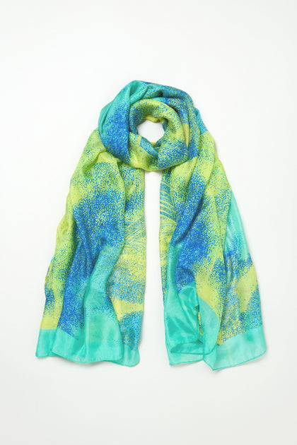 The Sky Dance Silk Scarf, featuring a blue, green, and yellow pattern and a fluid drape, is displayed flat on a white background.
