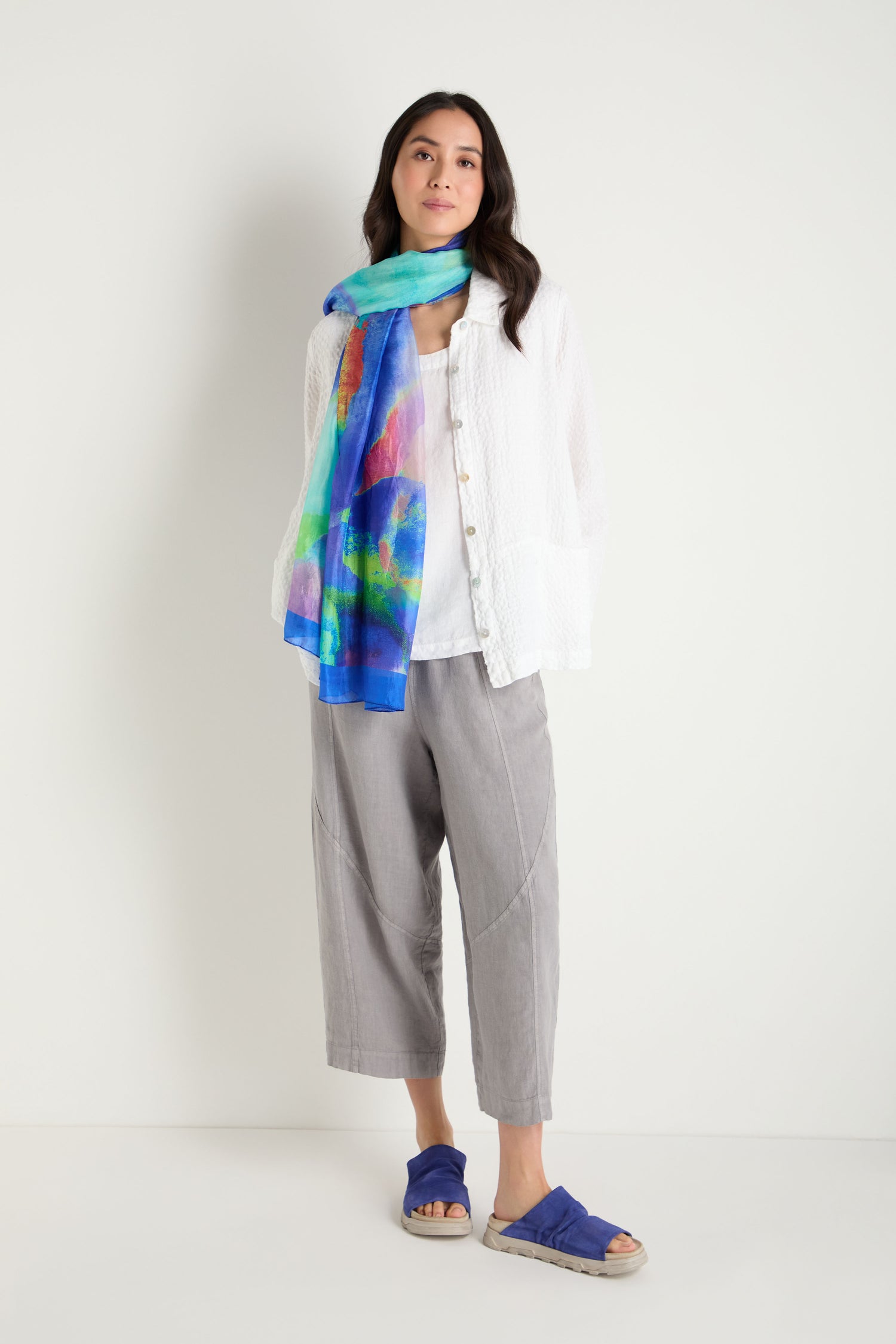 A woman stands against a plain background wearing a white textured top, gray cropped pants, the Aurora Veil Silk Scarf with an abstract print, and blue sandals.