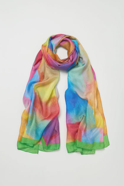 The Aurora Veil Silk Scarf, featuring an abstract multicolored watercolor print and green edges, is laid flat on a white background.