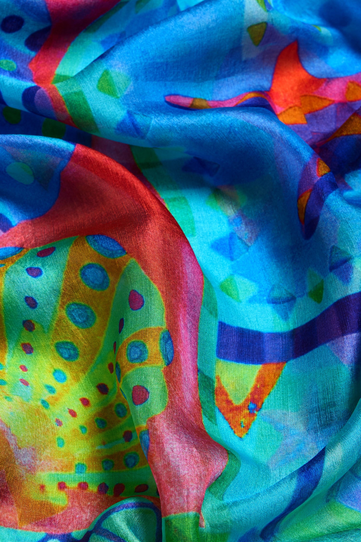 Close-up of the Ocean Garden Silk Scarf, showcasing its vibrant blue, red, green, and yellow abstract swirls and dots on lustrous silk—an elegant accessory with a luxurious sheen.
