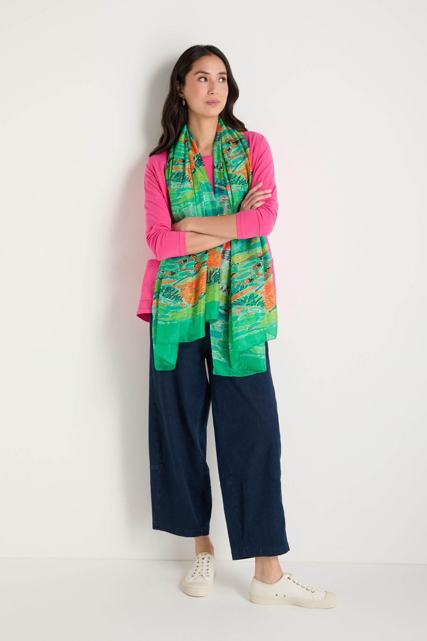A woman stands against a white wall wearing a pink top, blue wide-leg pants, white sneakers, and the Meadow Silk Scarf with her arms crossed.