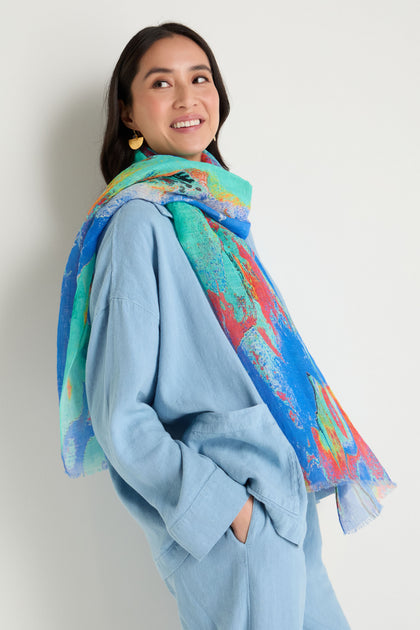 A woman smiles and looks to the side, standing with hands in pockets against a plain background, wearing a light blue outfit paired with the Terra Mare Cotton Linen Scarf.