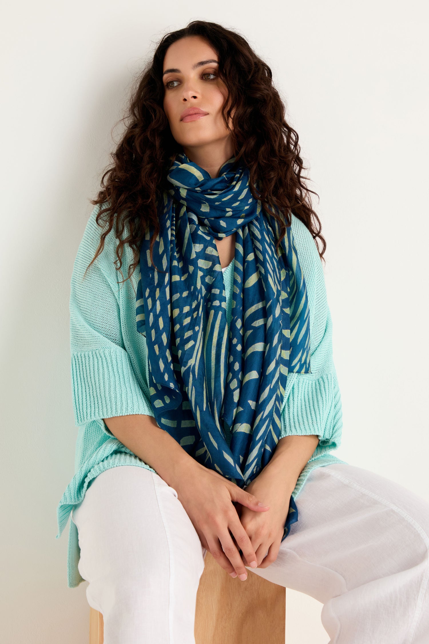 A woman with long curly hair wears a light blue sweater, white pants, and the Painted Waves Silk Scarf, sitting on a wooden stool against a plain light background.