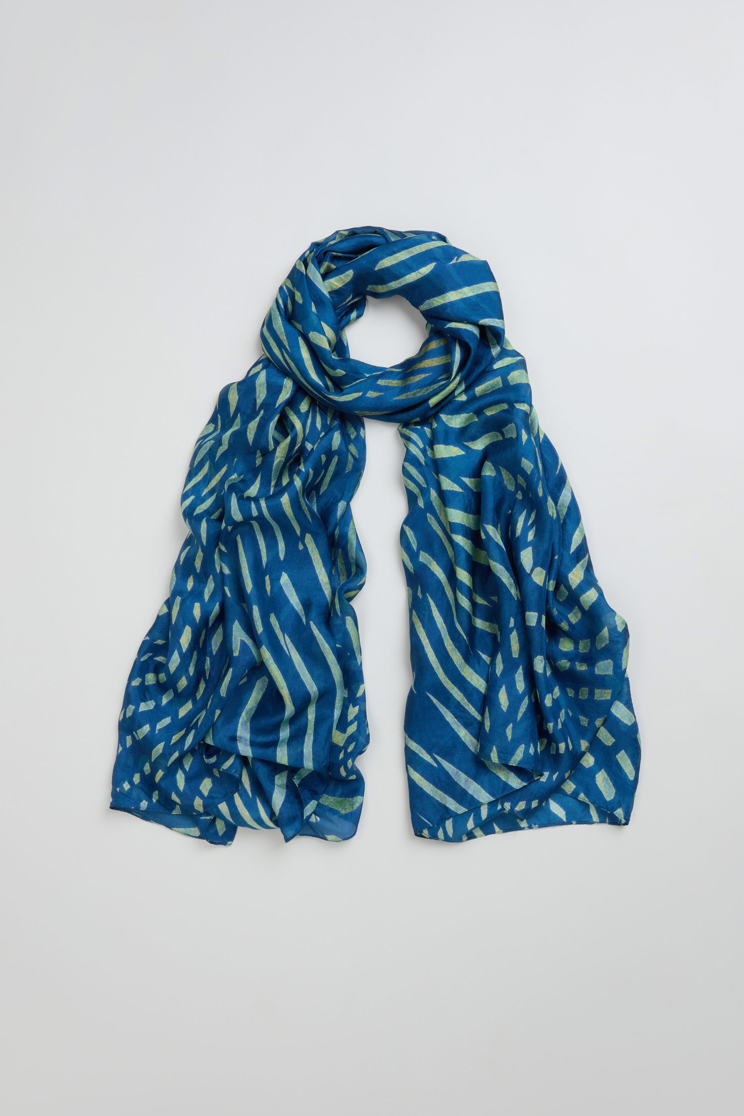 The Painted Waves Silk Scarf, featuring blue turquoise silk with light blue and white abstract diagonal and rectangular patterns, is displayed flat on a plain light background.