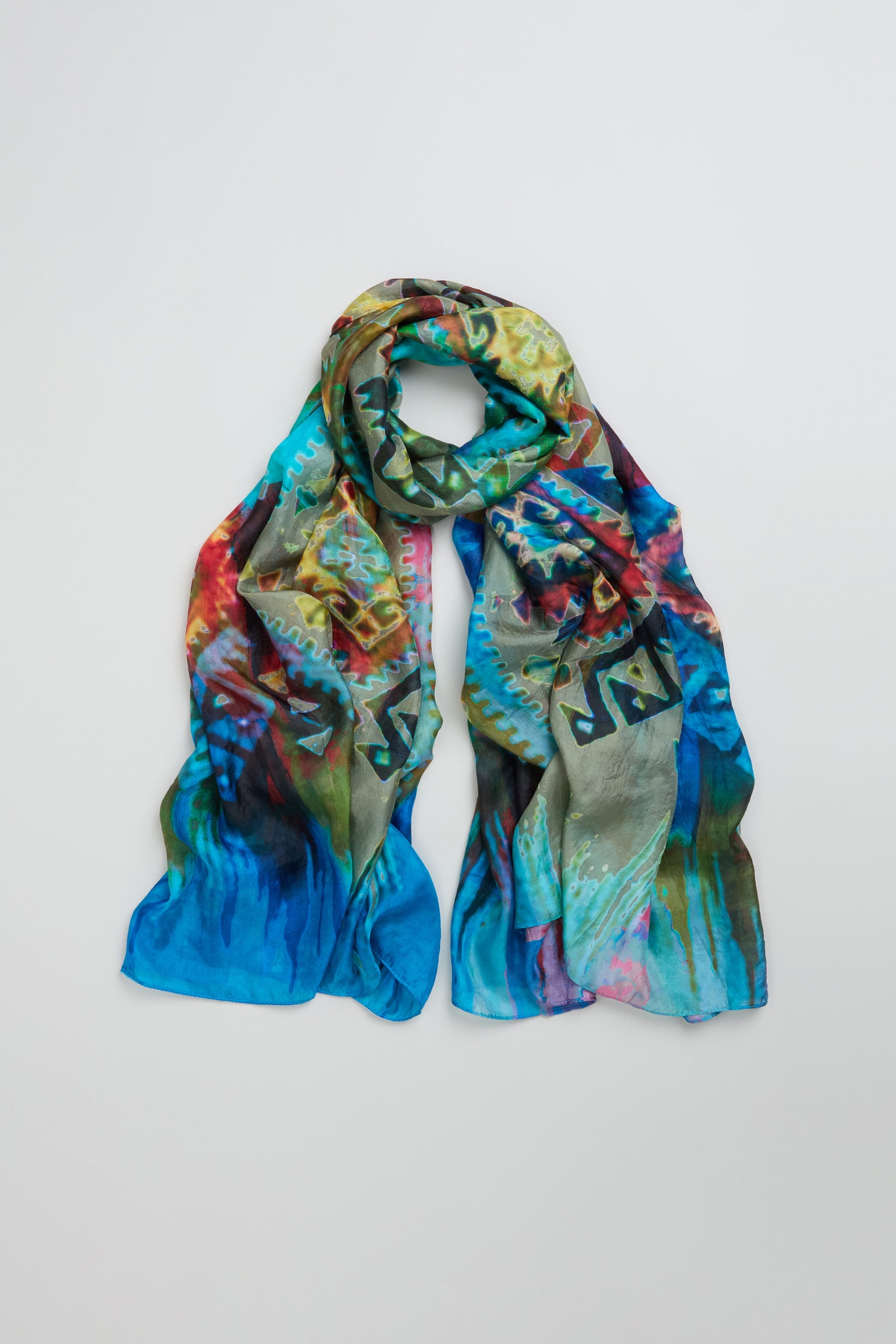 The Stained Glass Silk Scarf features colorful abstract patterns in blue, green, red, and yellow tones, arranged in a loose loop on a plain light background.