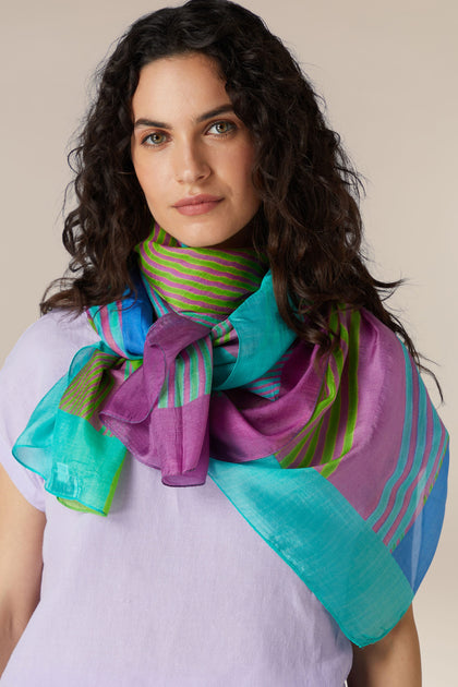 A woman with curly hair wearing a pastel purple top and a Candy Stripe Silk Scarf, adding a luxurious touch to her summer wardrobe.