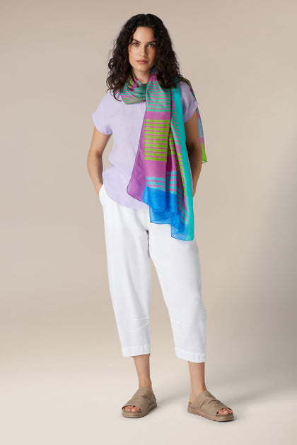 Woman standing in a relaxed pose wearing white capri pants, a lilac t-shirt, and a luxurious Golden Ray Silk Scarf, with brown sandals.