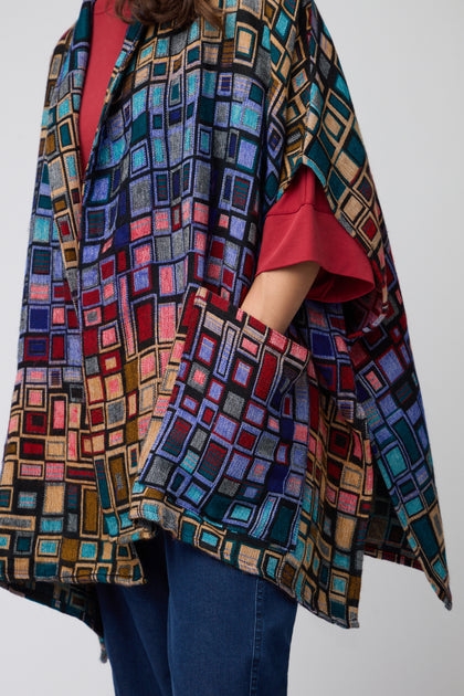 Model wears the Tesserae Jacket, featuring a colorful geometric pattern, over a red shirt and blue jeans, posed against a plain background.