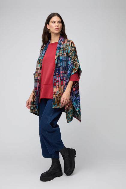 A woman stands against a plain background in a red top, blue jeans, black boots, and the Tesserae Jacket—a multi-tonal jacket featuring a colorful geometric pattern—exuding effortless style.