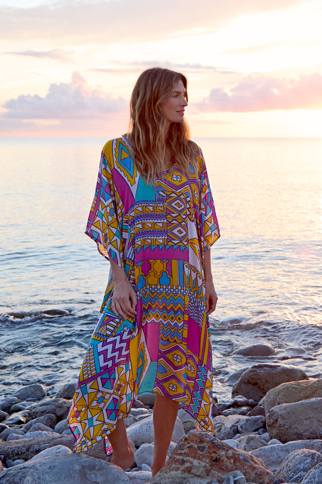 As the sun sets over a rocky beach, a person stands wrapped in an Elan V Neck Viscose Satin Kaftan. The vibrant geometric design enhances the tranquil scene of sky and sea adorned with gentle clouds.