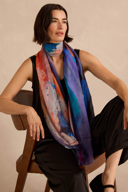 A person wears a Moontide Silk Scarf with an abstract watercolor pattern, draped over a black sleeveless top.