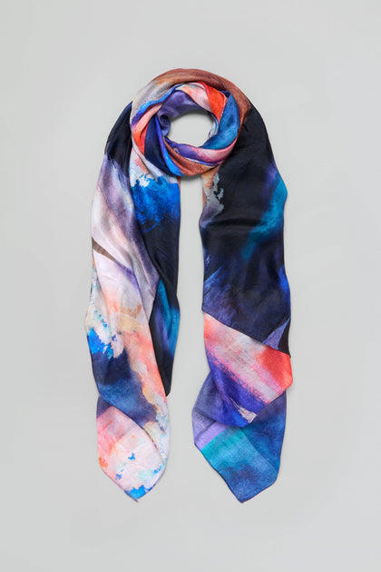 The Moontide Silk Scarf is a long, pure silk scarf featuring an abstract hand-painted print in shades of blue, orange, black, and white.