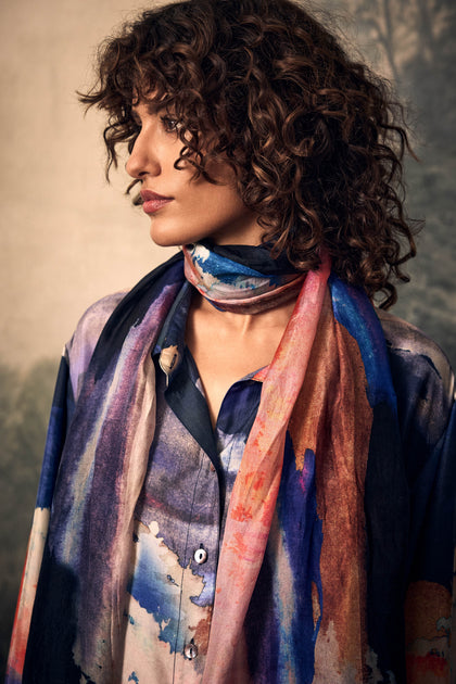A person wears a Moontide Silk Scarf with a colorful, hand-painted abstract watercolor pattern over a matching button-up shirt.