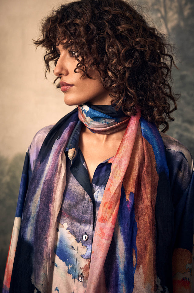 A person wears a Moontide Silk Scarf with a colorful, hand-painted abstract watercolor pattern over a matching button-up shirt.