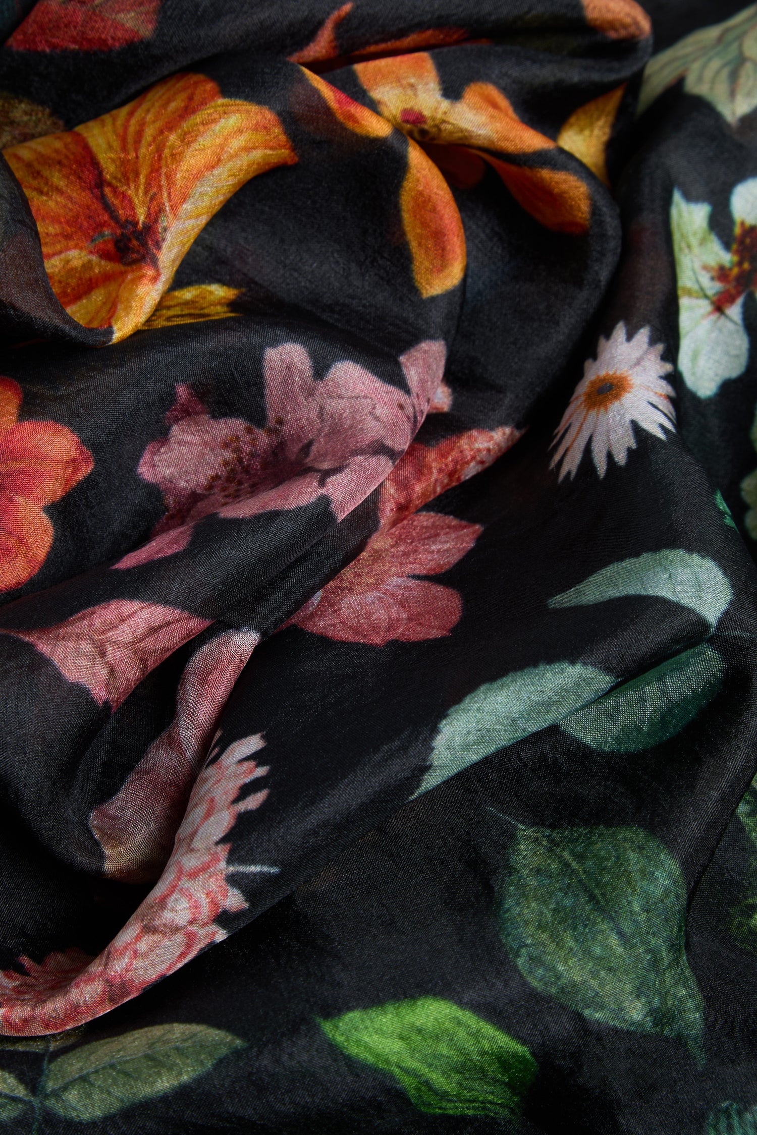 A close-up of the Midnight Garden Silk Scarf showcases its black fabric adorned with a vibrant botanical print, featuring orange, pink, and white flowers intertwined with green leaves.